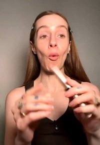 Jonge vrouw met lang haar houdt een make-upkwastje dicht bij haar gezicht, draagt een mouwloos topje tegen een effen achtergrond.