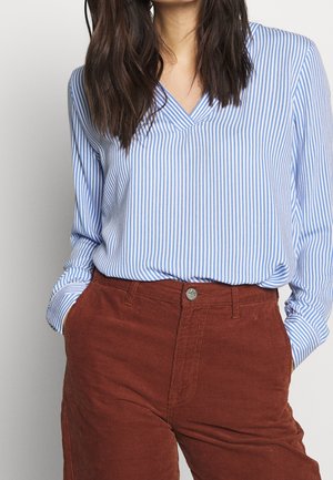 Femme portant un chemisier à col en V à rayures verticales bleu et blanc, rentré dans un pantalon en velours côtelé rouille taille haute, les mains dans les poches.