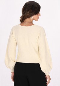 Cremfarbener Pullover mit Puffärmel und rundem Halsausschnitt. Gerippter Saum an den Bündchen und dem unteren Rand. Kombiniert mit schwarzen Hosen.