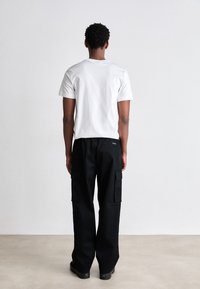 Hvid kortærmet t-shirt og sorte cargo-bukser med flere lommer, stående figur set fra ryggen mod en neutral baggrund.