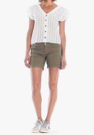 Mujer con blusa blanca de manga corta abotonada, pantalones cortos verdes oliva y zapatillas blancas con detalle de estrella, posando frente a un fondo blanco liso.
