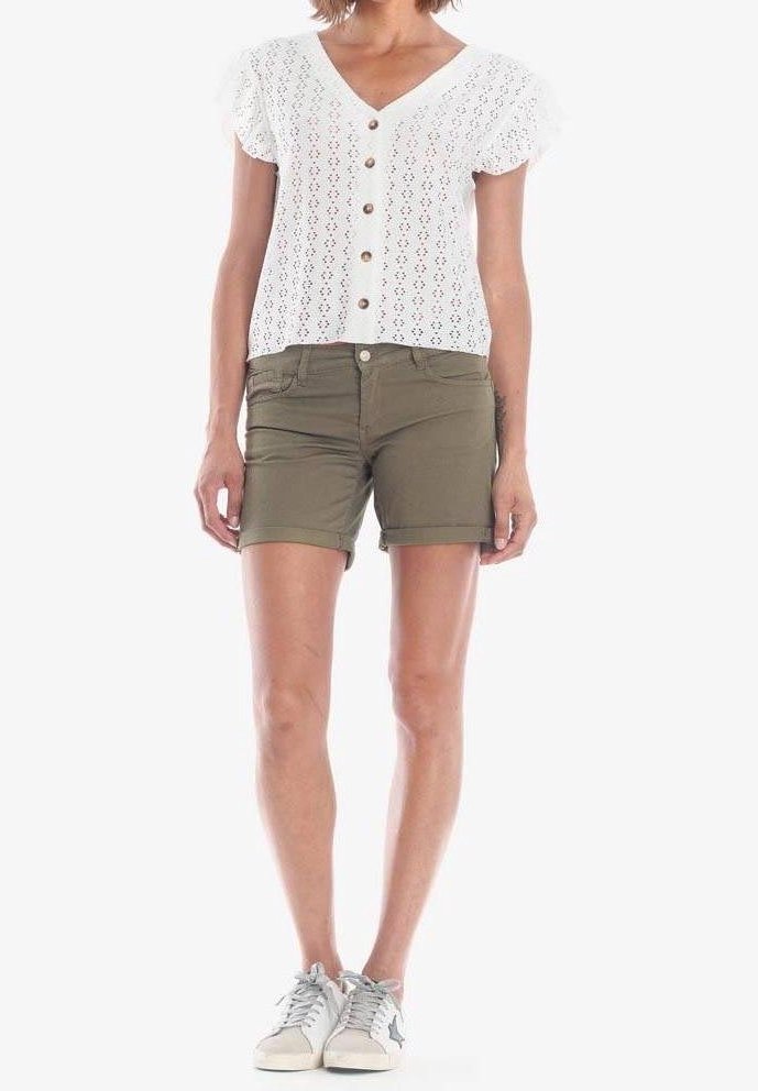 Femme portant un haut blanc à manches courtes boutonné, un short vert olive et des baskets blanches avec un détail étoile, debout devant un fond blanc uni.