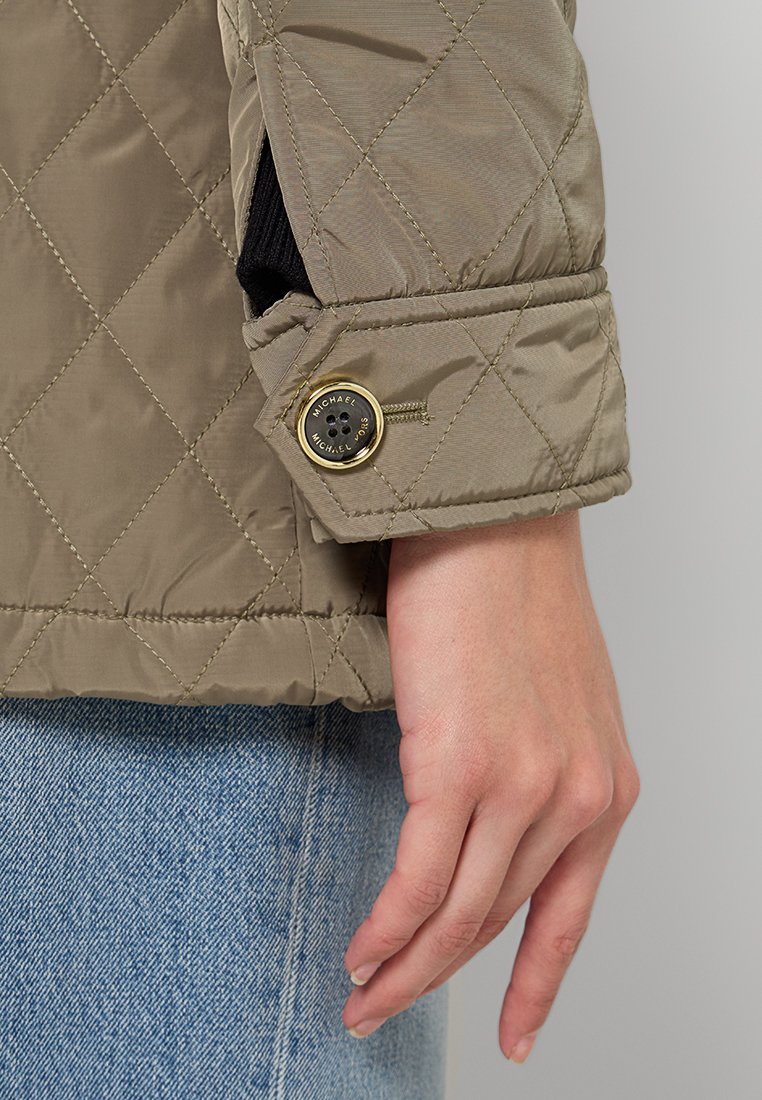 Gestepptes taupefarbenes Jacket mit glatter, glänzender Textur, versehen mit einem goldenen Akzentknopf und schwarzem Innenfutter. Hand ruht auf Denim-Stoff.
