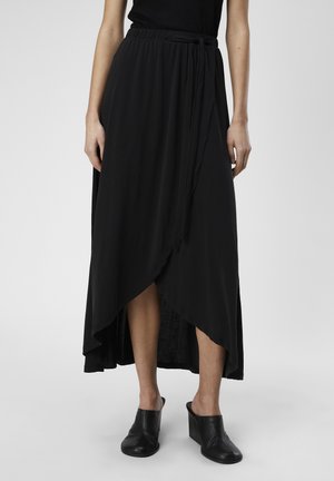 Jupe maxi noire en tissu léger et doux, avec un design enveloppant, un ourlet asymétrique et une ceinture à nouer à l'avant. Associée à des mules compensées noires.