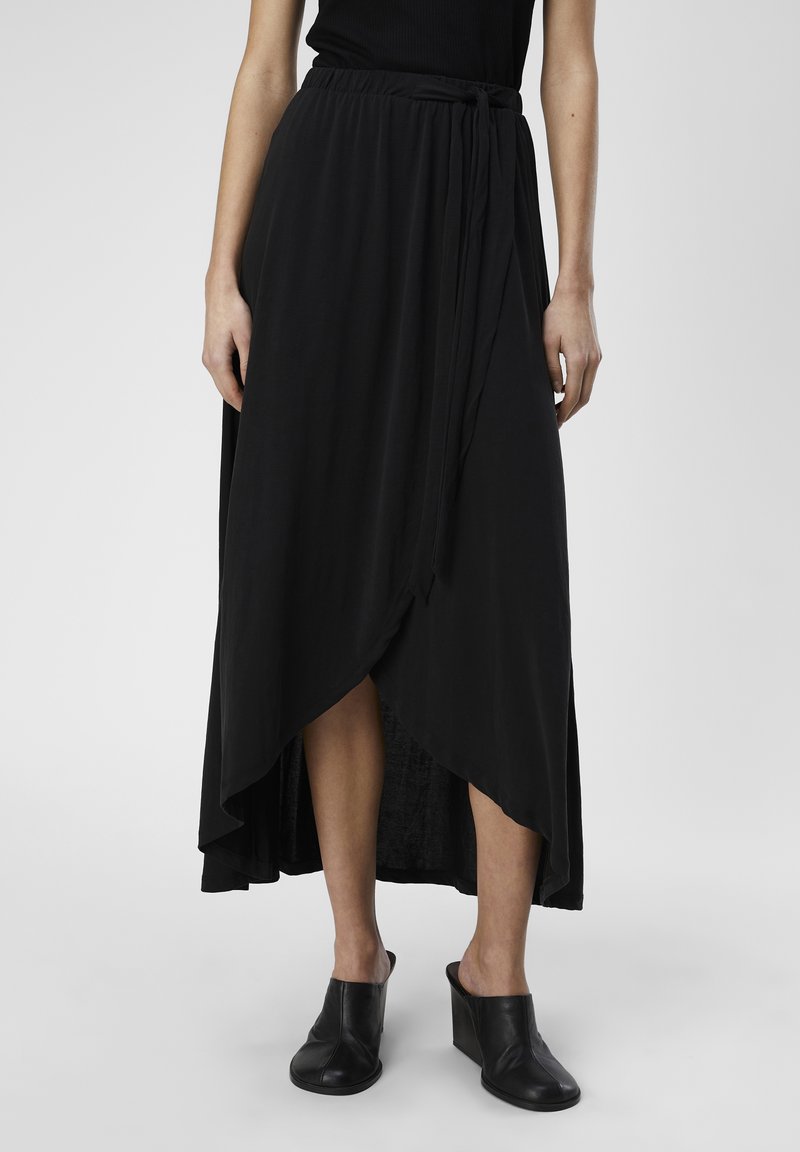 Jupe maxi noire en tissu léger et doux, avec un design enveloppant, un ourlet asymétrique et une ceinture à nouer à l'avant. Associée à des mules compensées noires.
