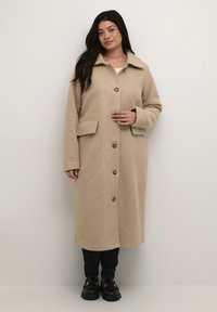 Beige fleecejakke med krave, lange ærmer, frontlukning med knapper og to sidelommer. Bæres over mørke bukser og sko.