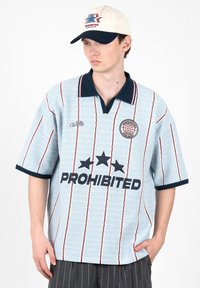 Jeune homme portant un polo rayé bleu clair avec « PROHIBITED » et des étoiles, une casquette blanche et marine, et un pantalon sombre à fines rayures, debout, la main dans la poche.