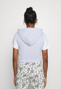 Sweat à capuche court bleu clair avec une fermeture éclair frontale et une capuche à cordon, associé à un pantalon blanc imprimé de motifs floraux avec des motifs bleus complexes.