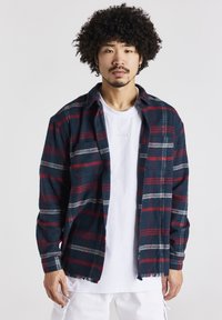 Camicia di flanella a quadri in blu scuro, rosso e bianco; con bottoni, colletto e orlo sfilacciato. Indossata su una maglietta bianca con dettaglio del logo.