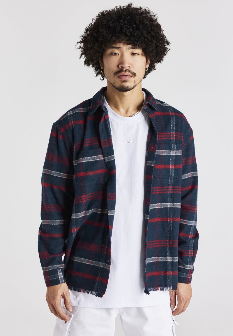Camicia di flanella a quadri in blu scuro, rosso e bianco; con bottoni, colletto e orlo sfilacciato. Indossata su una maglietta bianca con dettaglio del logo.