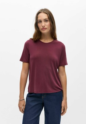 Vrouw met lichtbruin haar, die een bordeauxkleurig T-shirt met korte mouwen en een blauwe spijkerbroek draagt, staand tegen een effen witte achtergrond.