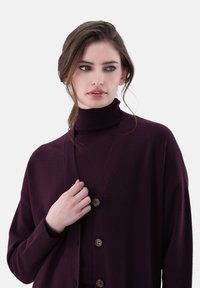 Burgunderfarbener Cardigan mit V-Ausschnitt und Holzknöpfen, aus weichem Material hergestellt, mit lockerem Schnitt und langen Ärmeln, über einem Rollkragenpullover getragen.