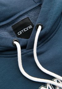 Sweat à capuche bleu avec cordons blancs, œillets métalliques noirs, et une étiquette noire à l'intérieur portant l'inscription "ombre."