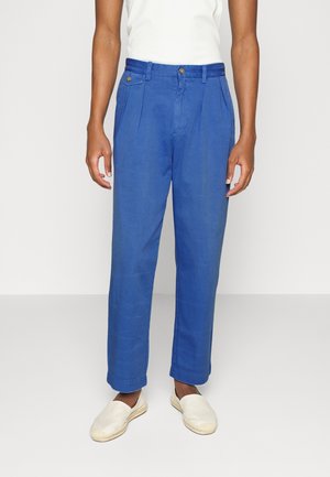 Pantalon en lin bleu avec plis, doté d'une fermeture à bouton et de poches latérales. Associé à des espadrilles de couleur claire.