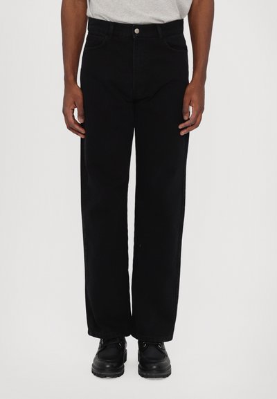 Pantalones de mezclilla negros de corte recto, con trabillas para cinturón, bolsillos delanteros y cierre de botón. Llevados con zapatos negros.