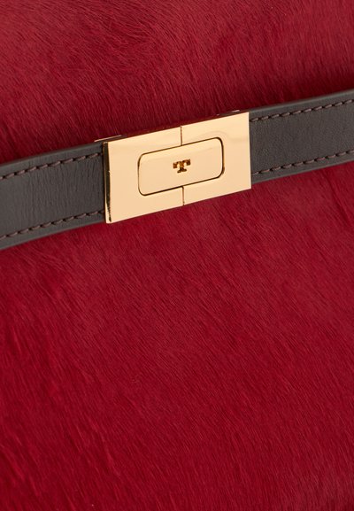 Surface texturée rouge avec une lanière horizontale en cuir marron foncé et un fermoir métallique doré rectangulaire au centre.