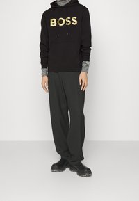 Felpa nera con il testo dorato "BOSS", indossata sopra una maglietta a righe grigia a maniche lunghe. Completata da pantaloni neri larghi e robuste stivali neri.