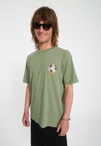Homme aux cheveux mi-longs portant des lunettes de soleil noires, un t-shirt vert olive avec un logo de globe, un pantalon noir et des bracelets, souriant à la caméra.