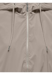 Veste beige avec une fermeture éclair argentée au centre et des cordons assortis munis d'embouts métalliques à la capuche.