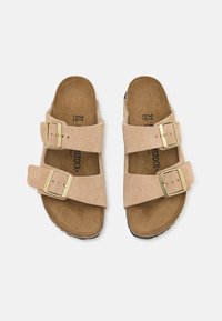 Sandales en suède beige clair avec deux brides réglables ornées de boucles en métal doré, semelle intérieure ergonomique et semelle extérieure noire durable.