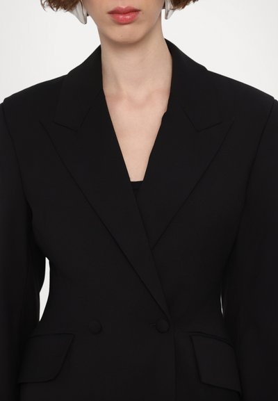Musta blazer, jossa on terävät silkot, kaksoispainonappi ja etupocketit. Sujuva kangas, muotoiltu istuvuus, minimaalinen muotoilu.