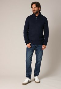 Pull marine avec un col zippé, associé à un jean bleu et des baskets blanches. Texture douce, design ajusté et style décontracté.