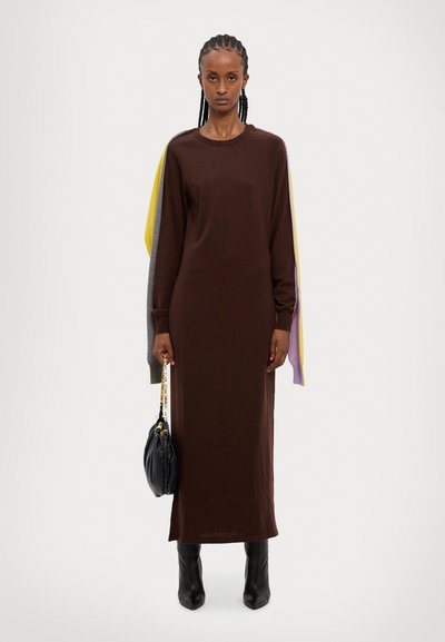 Robe longue marron à manches longues, avec des accents colorés en jaune, gris et violet. Portée avec un sac à épaule noir arrondi.