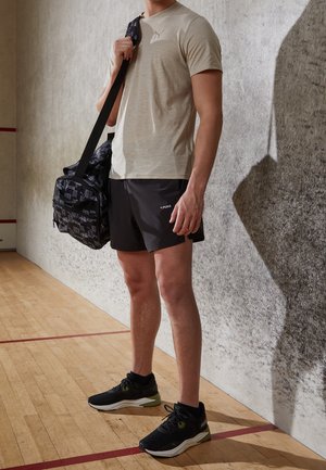 Chemise beige clair à manches courtes, short noir, baskets noires avec des accents verts, et un sac de sport noir à motif. Fond en plancher en bois.