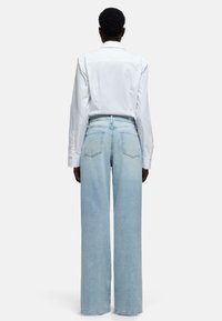 Camicia bianca a maniche lunghe abbinata a jeans a gamba larga di colore azzurro chiaro. I jeans presentano una chiusura con bottoni e due tasche posteriori con cuciture visibili.