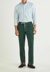 Camisa de botones a rayas en azul y blanco con puños naranjas, combinada con pantalones verdes con cordón y zapatos negros.