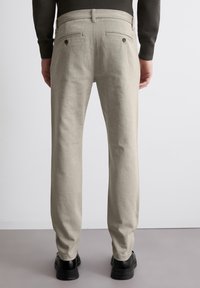 Helle beigefarbene Hose aus einem strukturierten Stoff, mit aufgesetzten Taschen und einem geraden Bein. Getragen mit schwarzen Schuhen.