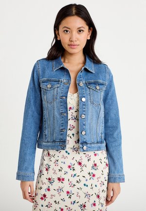 Kobieta ubrana w średnio niebieską kurtkę dżinsową na białą sukienkę w kwiaty z koronkowym dekoltem, stojąca na jednolitym białym tle.