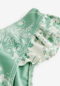 Zeleno cvetno kopalke imajo gladko tkanino z belim nabranim robom. Oblikovanje vključuje zapletene bele vzorce na svetlo zeleni podlagi.