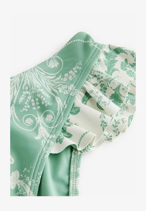 Le maillot de bain floral vert présente un tissu lisse avec une bordure froncée blanche. Le design comprend des motifs blancs complexes sur un fond vert clair.