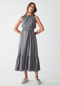 Vestido sin mangas gris con escote fruncido, cintura ceñida y volantes en capas en el dobladillo. Combinado con zapatos slip-on de rejilla blancos.