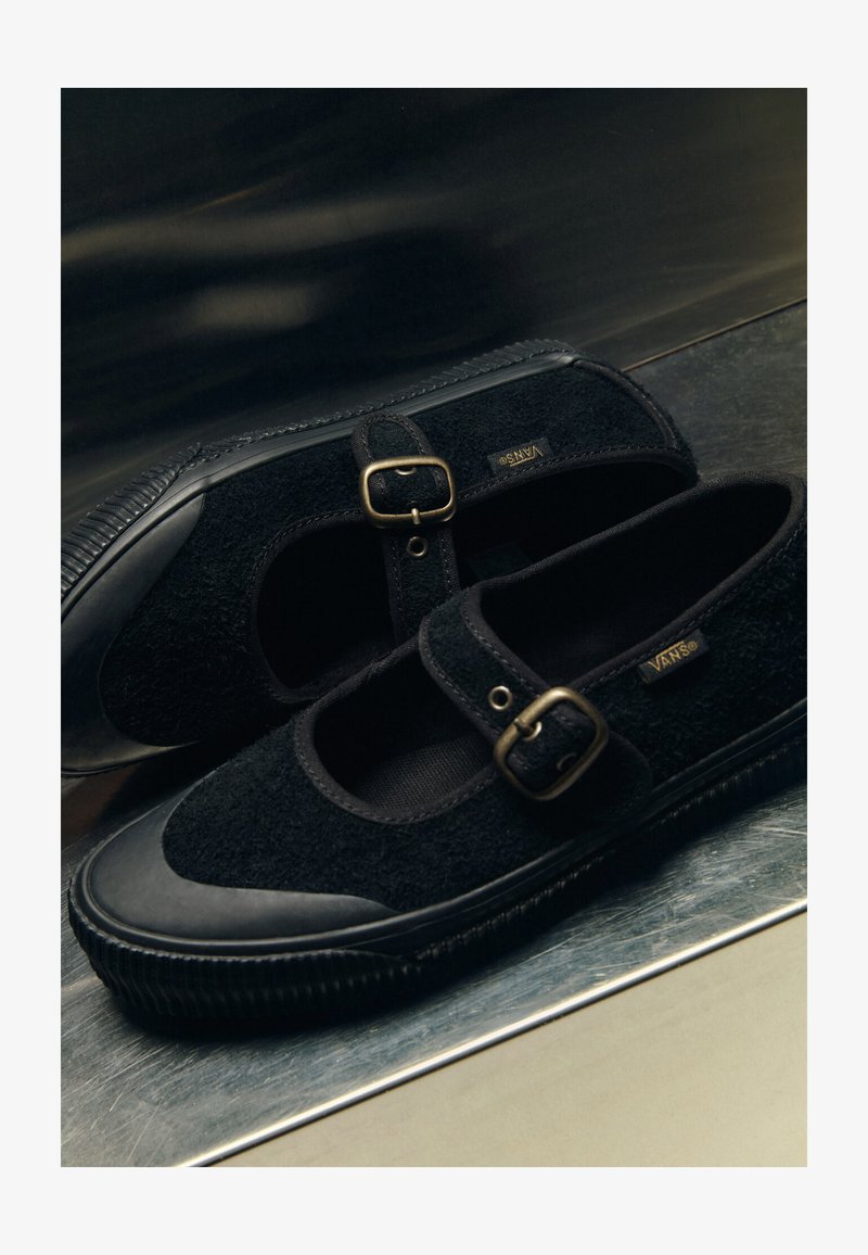 Paire de chaussures Vans Mary Jane noires avec tissu texturé, bouts en caoutchouc, semelles épaisses côtelées et brides à boucles en laiton sur une surface réfléchissante.