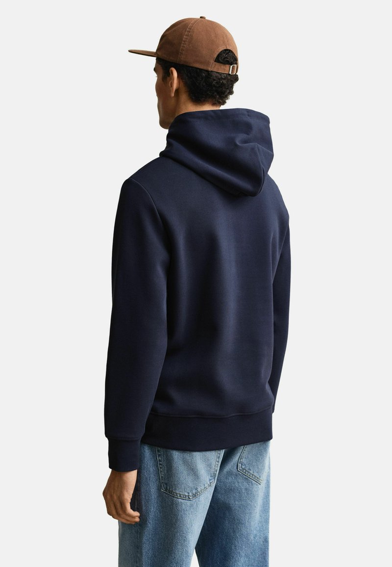Mann mit dem Rücken zur Kamera, trägt einen dunkelblauen Kapuzenpullover, hellblaue Jeans und eine braune Baseballkappe vor einem einfarbigen hellen Hintergrund.
