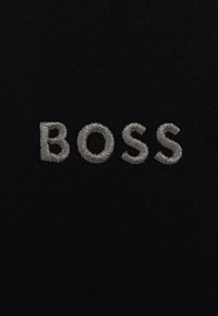 Crna tkanina s srebrnim izvezenim natpisom "BOSS" u jednostavnom, modernom fontu. Glatka tekstura s mat završetkom.