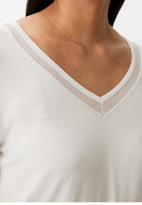 Sweter z dekoltem w serek w jasnym kremowym kolorze, z fakturowanym ściegiem i subtelnymi beżowymi akcentami wokół dekoltu, gładkim wykończeniem tkaniny oraz dopasowanym krojem.