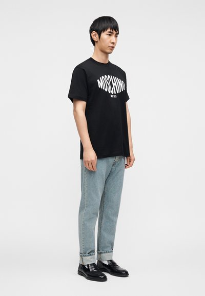 Μαύρο βαμβακερό T-shirt με το λογότυπο "MOSCHINO" τυπωμένο σε λευκό, συνδυασμένο με ανοιχτόχρωμα τζιν και μαύρα μοκασίνια, που στέκεται σε απλό φόντο.