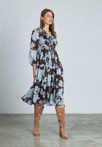 Vestido floral de longitud media con un estampado en marrón y azul claro, presenta un escote en V, detalles con volantes y mangas largas abullonadas. Combinado con botas marrones.