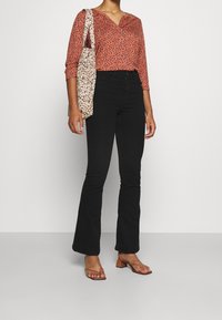 Blusa rosso-marrone con motivo floreale, scollatura a V, maniche a tre quarti; abbinata a jeans neri a campana. Accessoriata con una borsa a tracolla floreale.