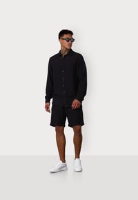 Schwarze langärmelige knopfüber Shirt, schwarze Shorts und weiße Sneakers. Sonnenbrillen runden das Outfit ab. Minimalistisches Design und modern geschnitten.