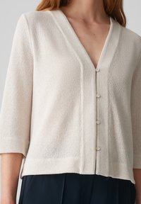 Hellbeige Cardigan mit V-Ausschnitt, drei viertel Ärmeln und geradem Saum, der sechs perlenähnliche Knöpfe auf der Vorderseite hat.
