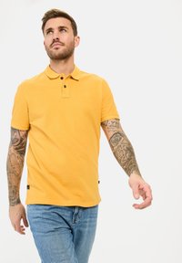 Geel poloshirt met een klassieke kraag, twee zwarte knopen en een klein reliëf logo. G gedragen met blauwe spijkerbroeken.