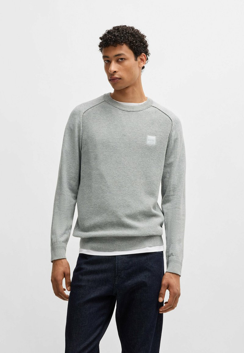 Helles graues Strick-Sweatshirt mit geripptem Rundhalsausschnitt, Raglanärmeln und einem kleinen Logopatch. Getragen über einem weißen T-Shirt, kombiniert mit dunkler Denim.