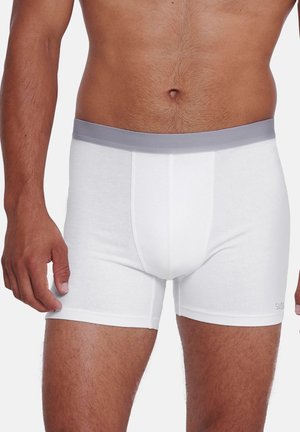 Mann trägt weiße Boxershorts mit grauem Bund, steht vor einem schlichten Hintergrund und zeigt die Körpermitte sowie die oberen Beine.