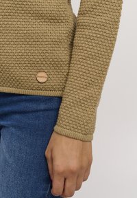 Beige knitted sweater with a textured finish, featuring a round wooden logo tag and ribbed cuffs, paired with blue denim jeans.
