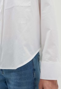 Camicia bianca button-down in tessuto leggero, con una tasca sul petto e un logo ricamato, abbinata a jeans in denim blu.