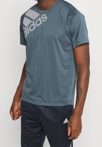 Man som bär en blå Adidas t-shirt med stort logo på bröstet och mörkblå Adidas träningsbyxor med logo på vänster lår.